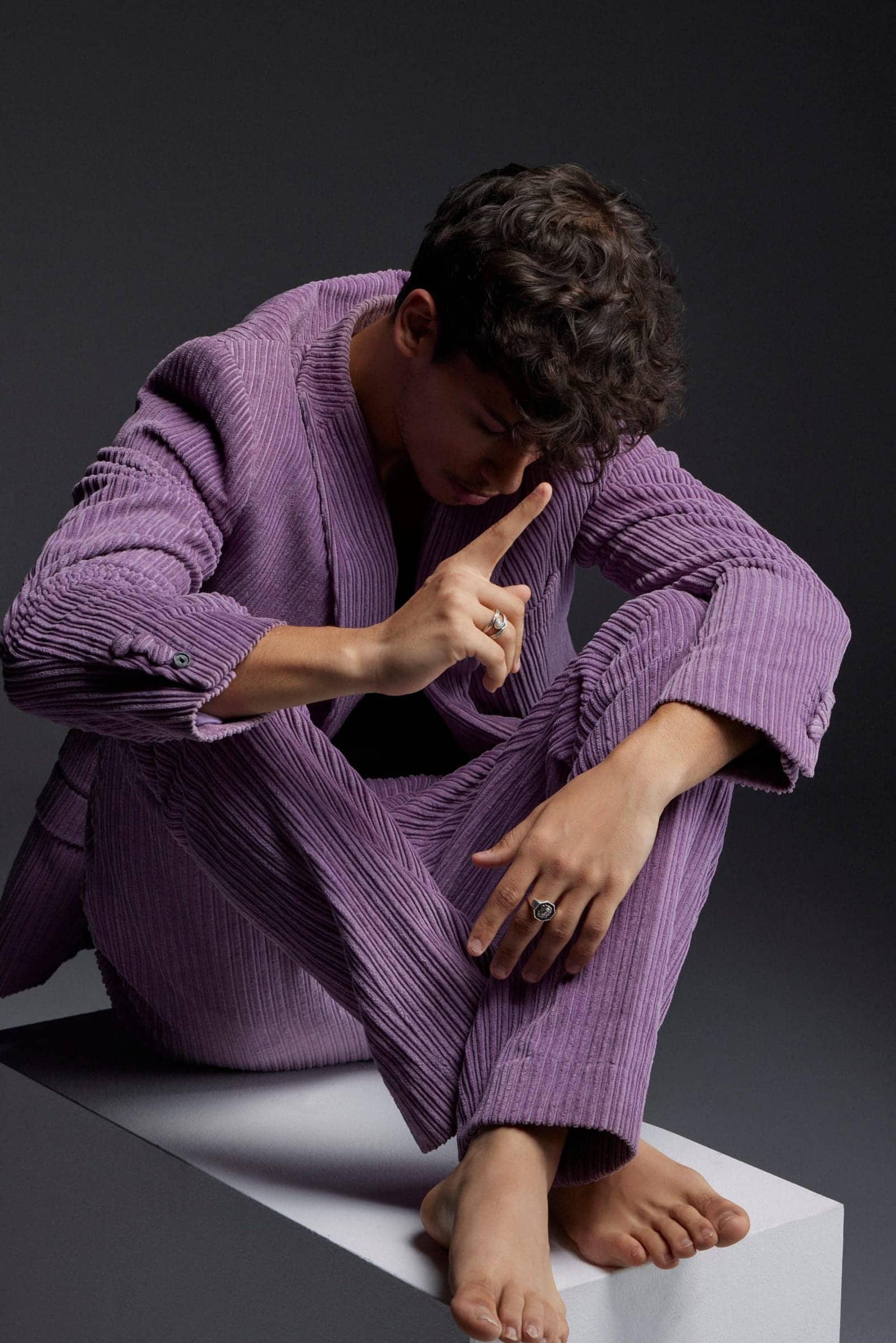 Harris J seated barefoot on a white block in a lilac corduroy suit