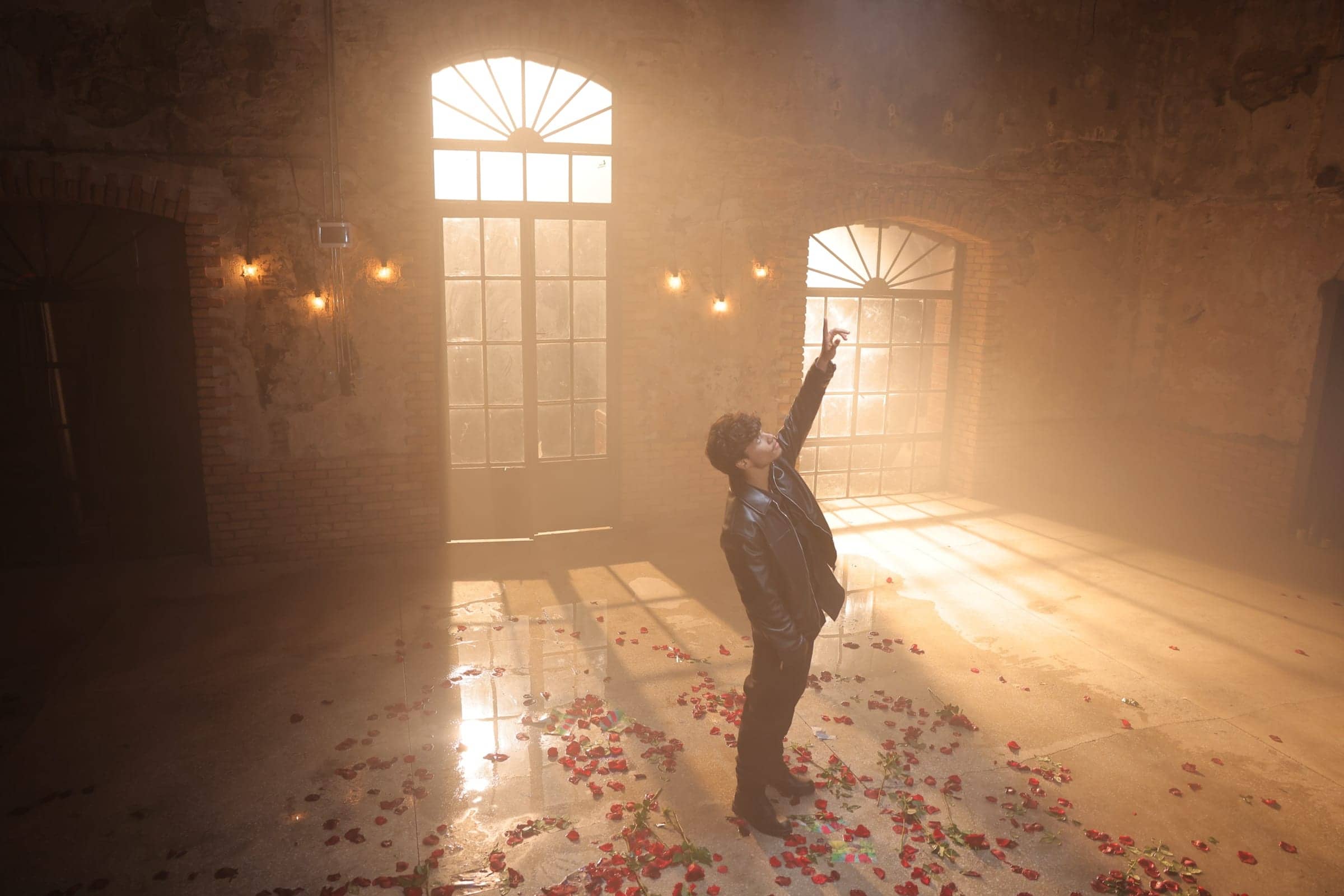 Harris J reaching upwards in a sunlit brick hall scattered with rose petals